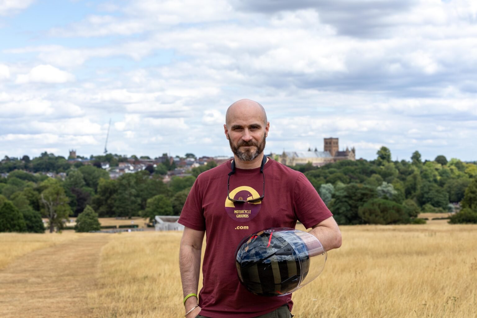Rider in a Motorcycle Grounds t-shirt holding a helmet in a field, with the St Albans skyline behind