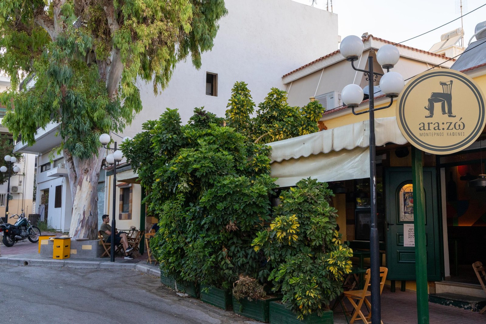 The exterior of Ara:zo in a residential Rhodes Town street, with the round logo showing a chair with crossed legs and a motorcycle parked nearby