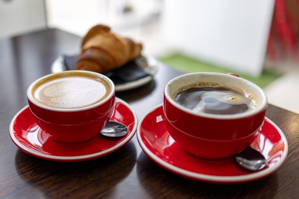 Two red cups of coffee on red saucers, with a croissant on a table behind