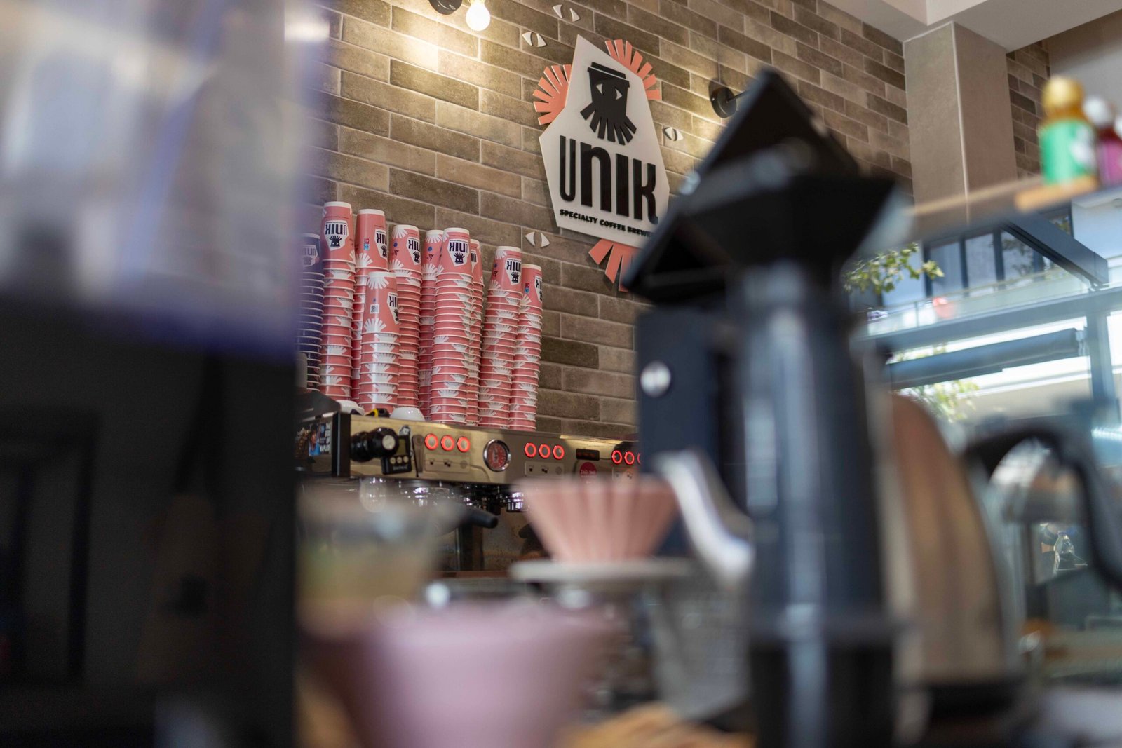 Behind the counter at Unik Specialty Coffee Brewers with the squid logo, stacked branded cups, and a pink pour-over dripper