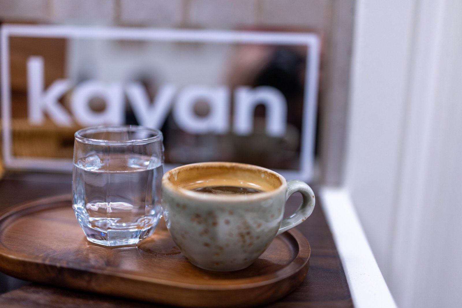 An americano served with a glass of water on a wooden tray, with the Kayan logo visible in the window behind