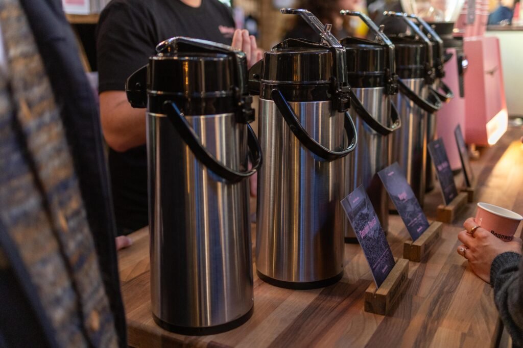 Row of stainless steel brew flasks with tasting notes on a wooden counter at the Beanworks stand
