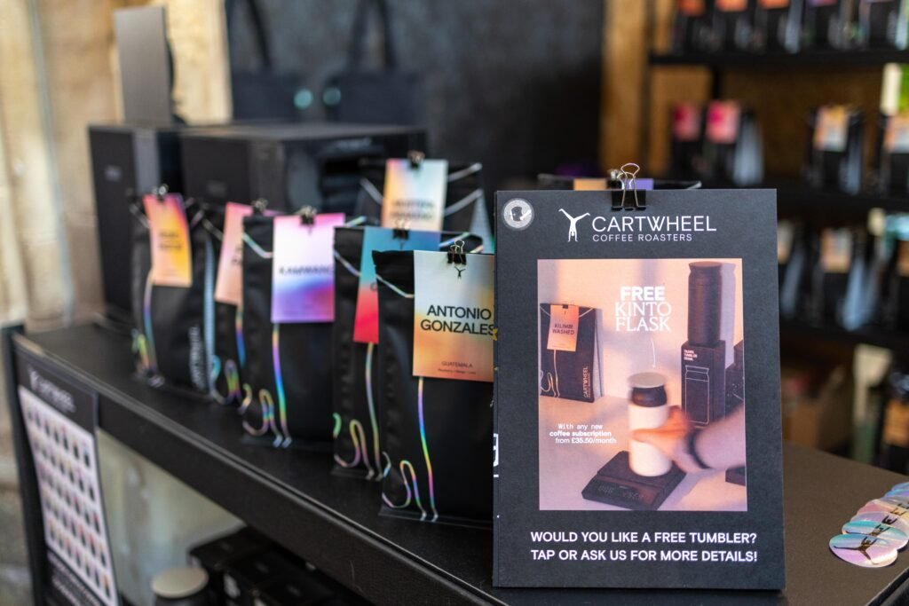Bags of Cartwheel Coffee Roasters beans with colourful tags lined up on the stand, beside a sign offering a free tumbler