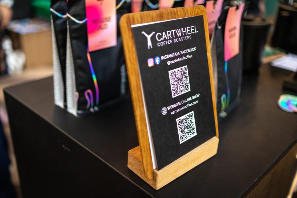 A wooden stand holding a Cartwheel Coffee Roasters card with social media QR codes, in front of coffee bags