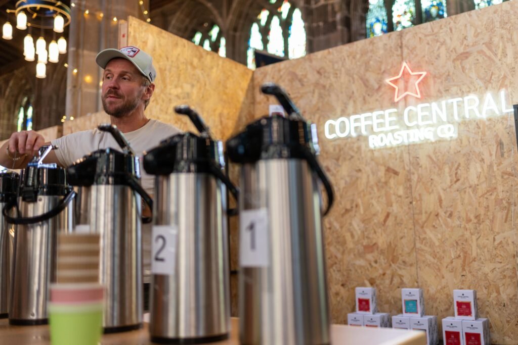 A Coffee Central team member in a cap behind a row of numbered airpot flasks, beside the neon sign