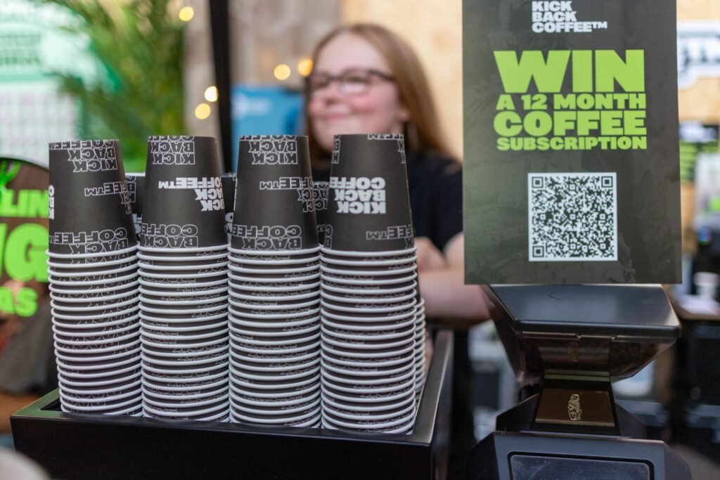 Stacks of Kickback Coffee paper cups beside a sign offering a 12 month coffee subscription