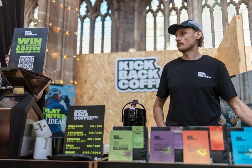 A Kickback Coffee team member in a black T-shirt and cap behind the counter, with coffee bags and a subscription sign