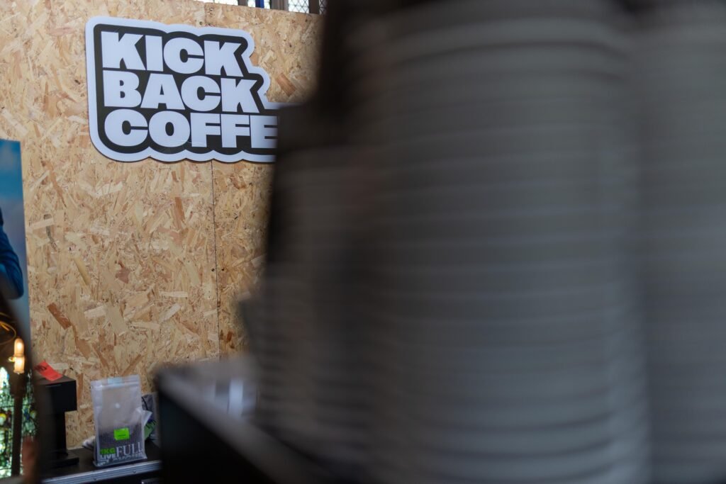 The Kickback Coffee logo on the wooden board, with a stack of cups blurred in the foreground