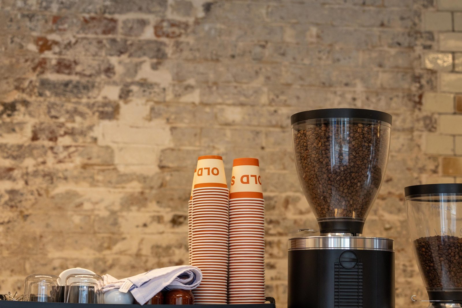 Stacked takeaway cups and a grinder full of coffee beans against a whitewashed brick wall