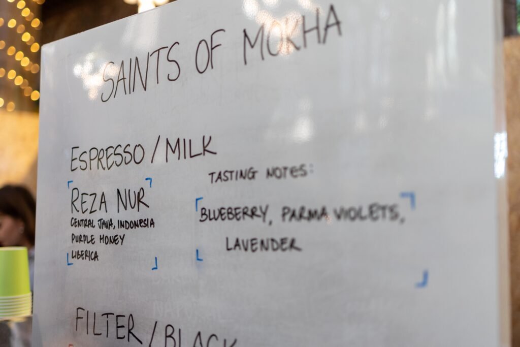 A whiteboard at the Saints of Mokha stand listing espresso and filter coffees with tasting notes