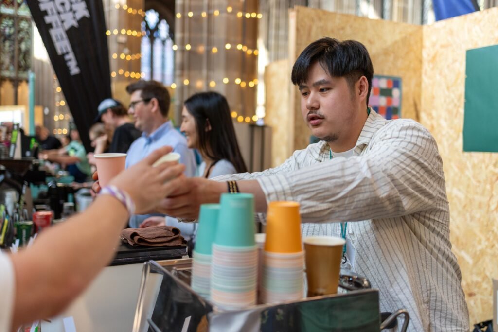 Man handing a coffee to a visitor at the Tilted Coffee stand, with teal and orange cups on the counter